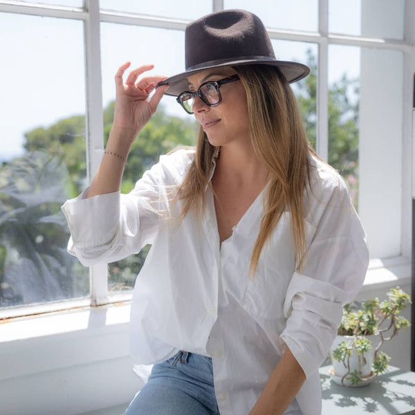 Brown wool felt fedora with leather trim, worn by a woman with long hair and glasses, complemented by a white shirt and jeans.