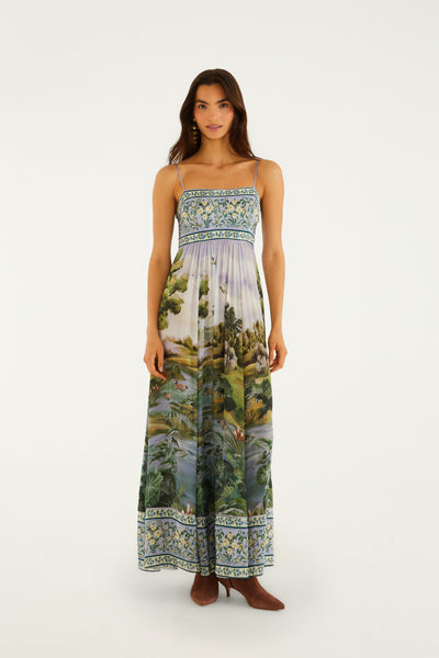 Maxi dress featuring a green landscape print with lavender skies, adjustable straps, and an ornamental border detail.