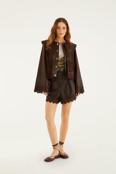 Brown leather blouse with intricate cut-out detailing, styled with matching shorts and flats. Model poses confidently.