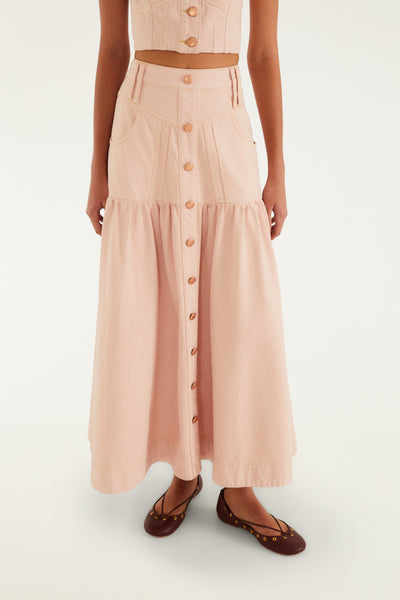 Pink cotton twill denim maxi skirt with a flared silhouette, button front, and tiered ruffle hemline. Worn with stylish flats.