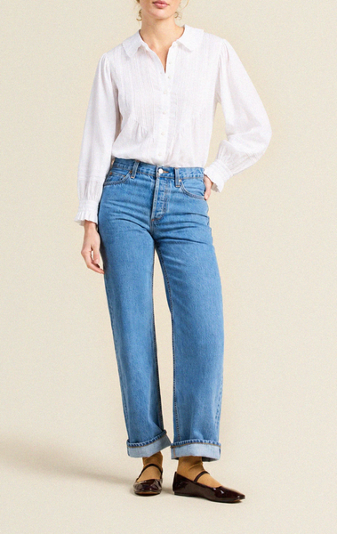 Fitted white cotton voile blouse with oversized point collar, pintucked front, and long sleeves with ruffled cuffs. Worn with high-waisted blue jeans.